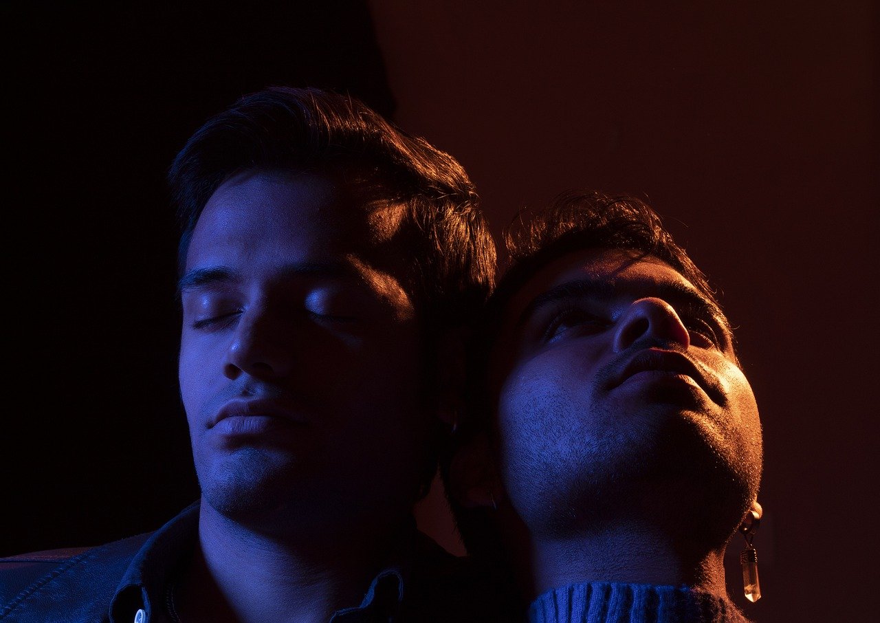 Deux jeunes hommes se tiennent l'un à côté de l'autre. Celui de gauche a les yeux fermés, tandis que celui de droite a la tête penchée en arrière, le regard vers le ciel.