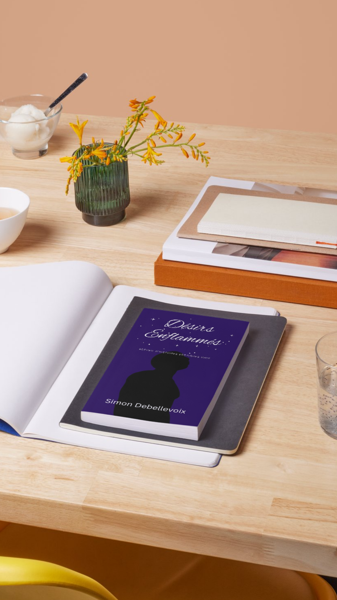 Désirs Enflammés : Recueil d'histoires érotiques gays Bureau en bois clair avec un livre violet intitulé « Désirs Enflammés » de Simon Debellevoix posé sur un cahier ouvert, entouré d’un carnet beige, d’un vase vert avec fleurs orange, d’un bol, d’un verre d’eau et d’un bol de sucre sur fond beige.