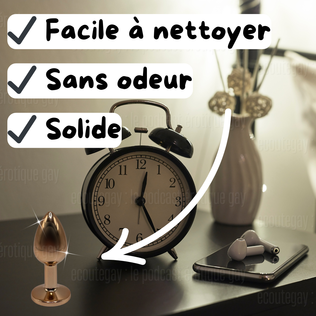 Table de nuit légèrement éclairée avec un vase composé de fausses fleurs, un smartphone, des écouteurs, un réveil à aiguilles et le fameux plug anal steel sont posés dessus.