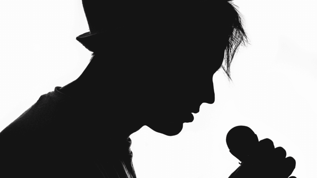 Silhouette noire élégante d'un jeune homme de profil, incliné vers un micro vintage comme pour murmurer des secrets intimes, fond blanc épuré. Symbole puissant de la voix, de la musique et de l'inspiration quotidienne à travers chansons et textes.