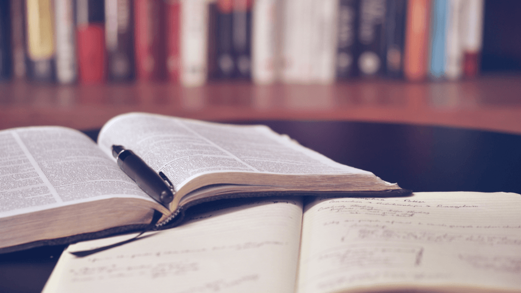 Deux livres ouverts sur une table en bois sombre, un stylo posé dessus, avec une bibliothèque floue en arrière-plan aux tons chauds orangés. Ambiance cosy et invitante pour l'étude et l'enrichissement personnel de l'esprit.
