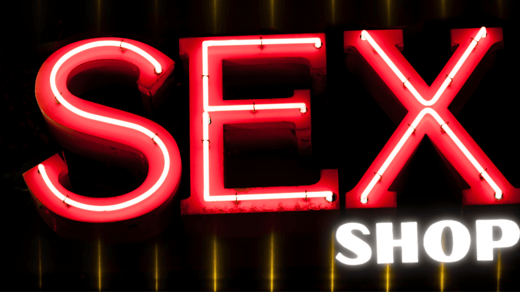 Enseigne néon iconique « SEX SHOP » en lettres rouges massives, illuminée avec lueur chaude orangée sur fond noir velouté, reflets métalliques brillants. Symbole torride du plaisir illimité et des achats érotiques. Partenariat pour podcast gay.