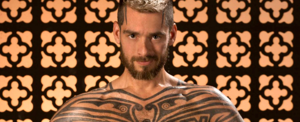 Logan McCree avec le torse tatoué, portrait en gros plan sur fond décoratif.