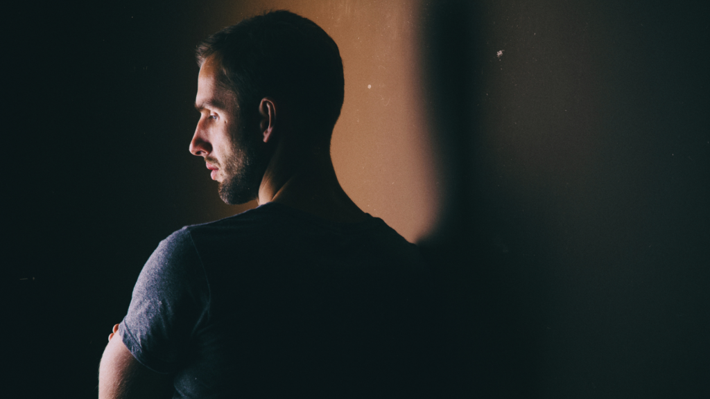 Homme pensif regardant au loin dans une pièce sombre, illustrant l'introspection et la mélancolie après une rencontre sexuelle.
